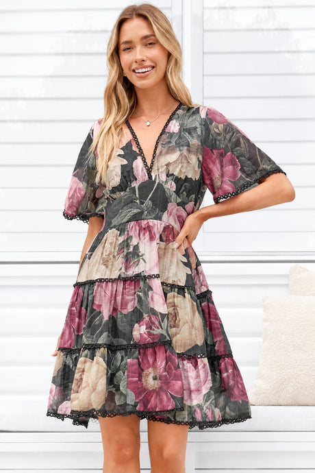A woman modeling a tiered floral dress with short flutter sleeves and black lace trim, standing in front of a white paneled wall.