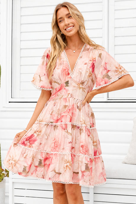 A woman models a light pink tiered mini dress with a floral print, featuring flutter sleeves and white lace trim, posed in front of a white shuttered window.