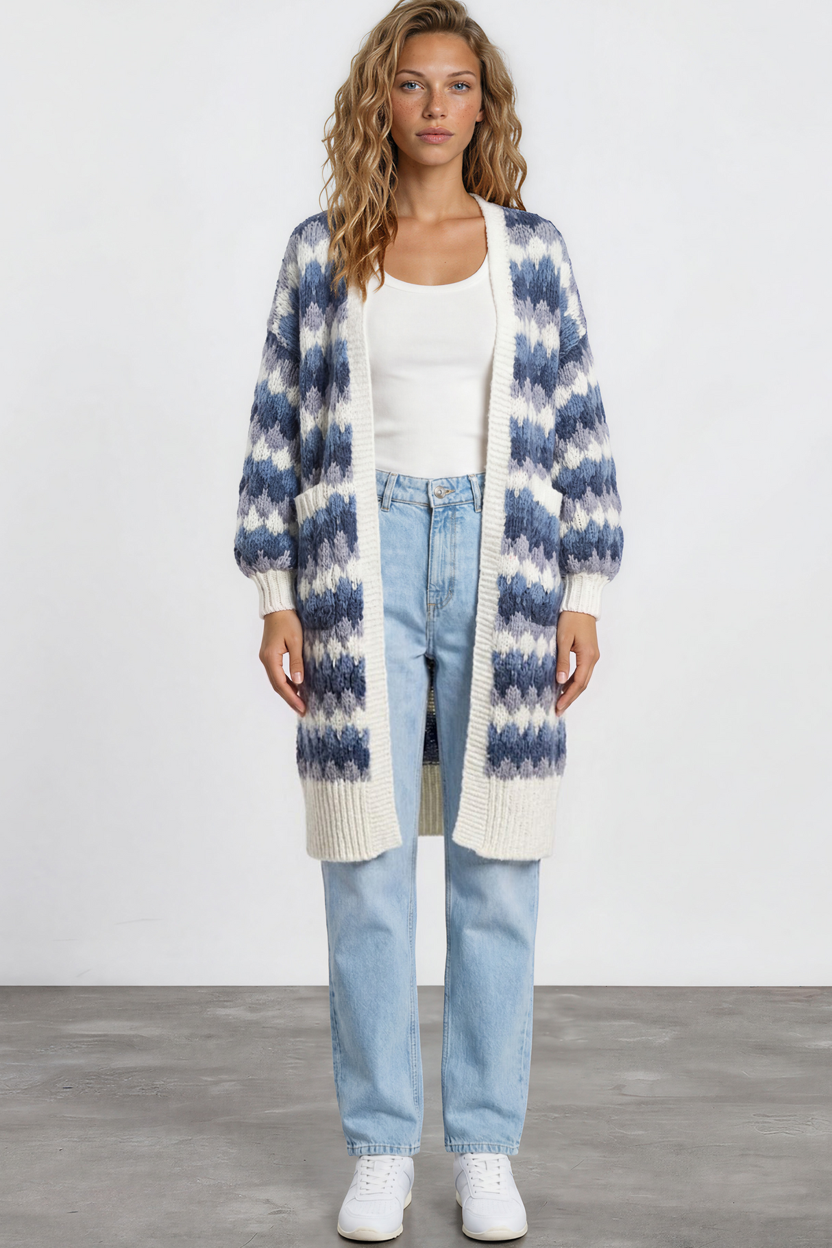 Woman wearing a long, patterned cardigan over a white top and jeans on a plain background