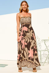 A smiling woman standing in the sunshine in the Mattina Maxi Dress in Ginevra Print, a spaghetti strap sun dress with a long A-line skirt in a brown, pink and tan tropical leaf print with a swirl border hem