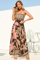 A woman with one hand in her hair and one on her hip wearing the Mattina Maxi Dress in Ginevra Print, a spaghetti strap sun dress with a straight neckline in a brown, pink and tan tropical leaf and swirl print, against a white wall