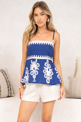 A woman standing with arms relaxed in the Meg Cami in Maceo Print Blue, a navy spaghetti-strap cami with a white trimmed bodice and white printed peplum, styled with white shorts against a light wall and striped cushions