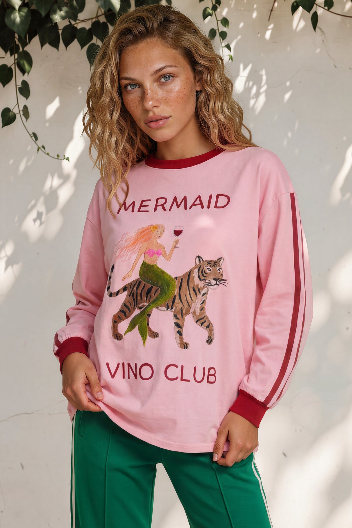 Woman wearing a pink 'Mermaid Vino Club' sweatshirt with a tiger graphic against a white wall.