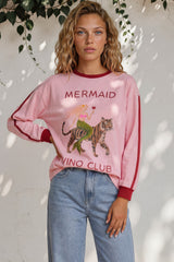Woman wearing a pink 'Mermaid Vino Club' sweatshirt with a tiger graphic, standing against a white wall with greenery.