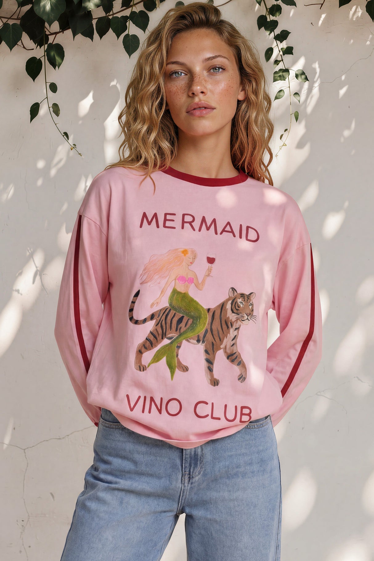 Woman wearing a pink 'Mermaid Vino Club' sweatshirt with a tiger graphic, standing against a white wall with greenery.