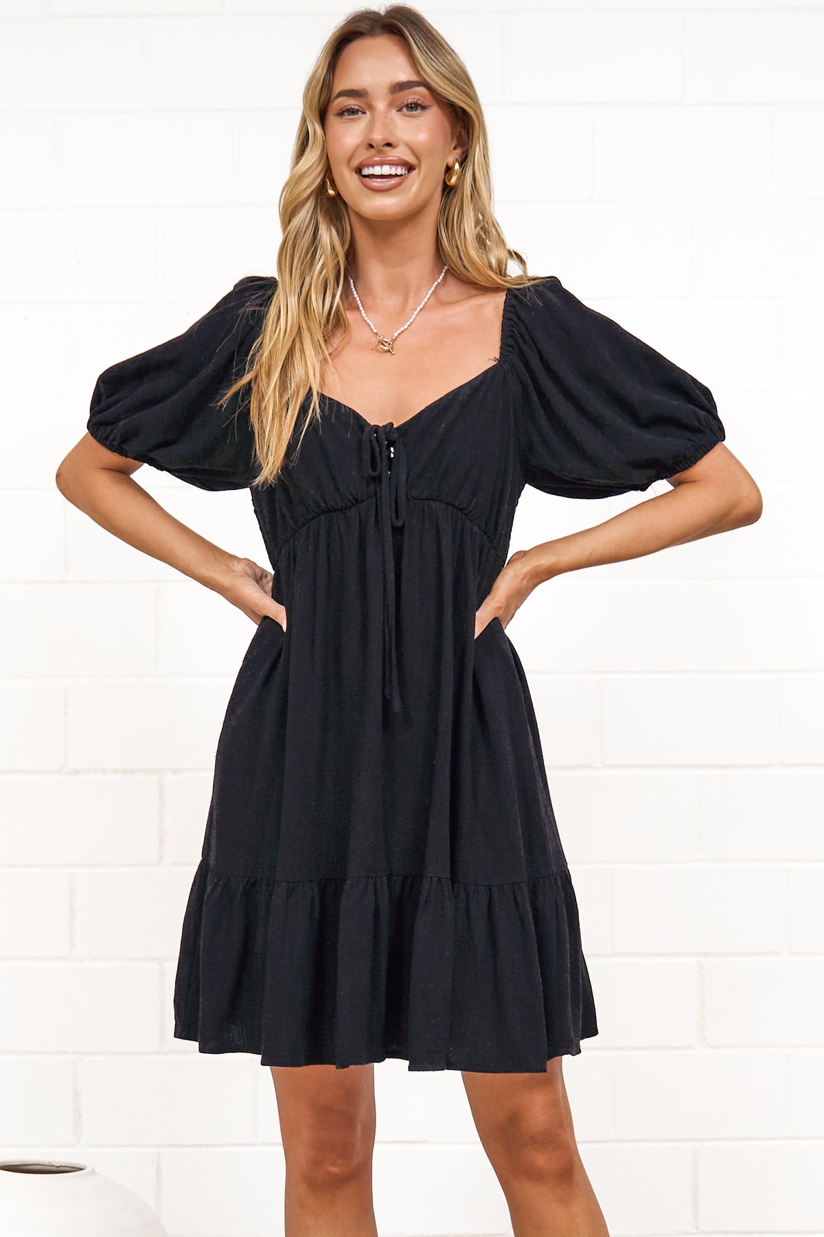 A woman in a black mini dress with a sweetheart neckline, tie-front detail, puff sleeves, and tiered skirt, posed against a white painted brick wall.