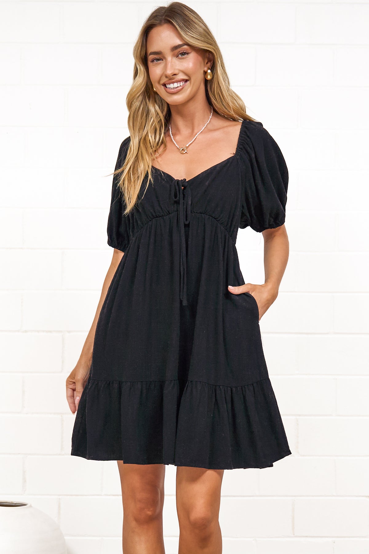 A woman in a black mini dress with a sweetheart neckline, tie-front detail, puff sleeves, and tiered skirt, posed against a white painted brick wall.