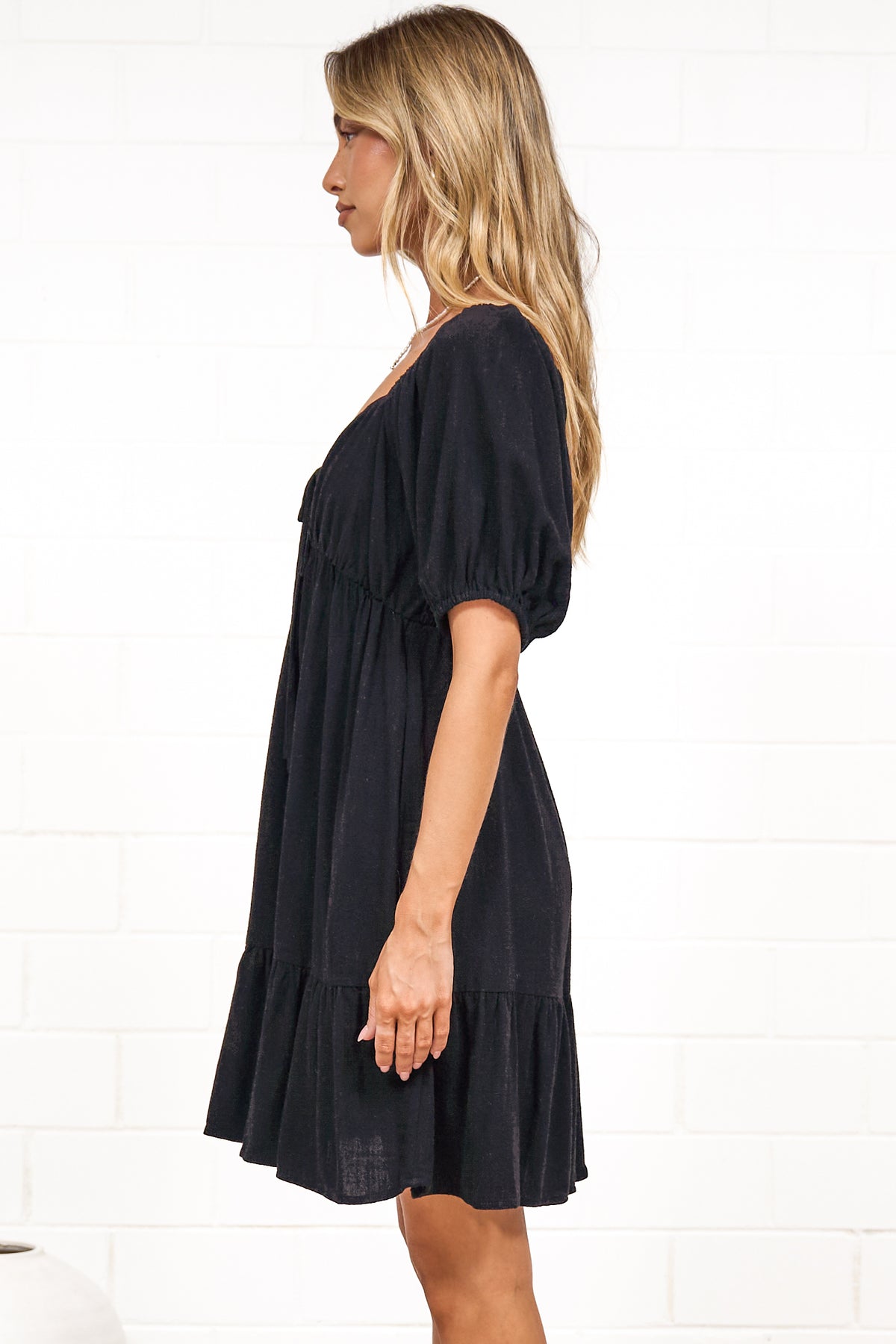 A woman in a black mini dress with a sweetheart neckline, tie-front detail, puff sleeves, and tiered skirt, posed against a white painted brick wall.