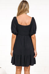 A woman in a black mini dress with a sweetheart neckline, tie-front detail, puff sleeves, and tiered skirt, posed against a white painted brick wall.