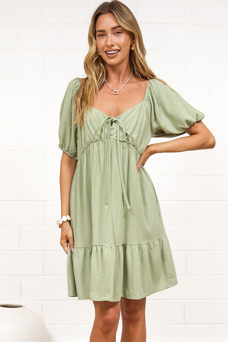 A woman wearing a sage mini dress with a sweetheart neckline, tie-front detail, puff sleeves, and tiered skirt, standing against a white painted brick wall.