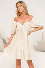 A woman modeling an off-the-shoulder cream-colored mini dress with puff sleeves, a tie-front detail, and a ruffled hem, set against a light-colored indoor background.