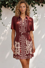 Woman wearing a patterned dress standing against a white wall with greenery.