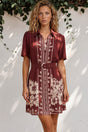 Woman wearing a patterned dress standing against a white wall with greenery.