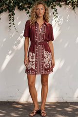 Woman wearing a patterned dress standing against a white wall with greenery.