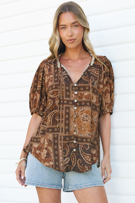A woman standing with arms relaxed in the Neo Blouse in Mahalla Print, showing the untucked relaxed fit, frill collar and button front of the brown paisley patchwork blouse, with light denim shorts against a white wall