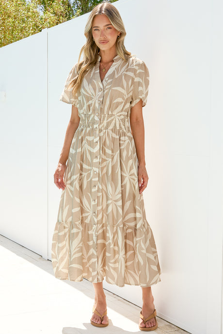 A woman wearing the Paloma Midi Dress in Gretchen Print, a beige and cream leaf-print A-line button-down midi with a split mandarin collar and cap sleeves, paired with tan sandals against a sunlit white wall