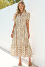 A woman wearing the Paloma Midi Dress in Gretchen Print, a beige and cream leaf-print A-line button-down midi with a split mandarin collar and cap sleeves, paired with tan sandals against a sunlit white wall