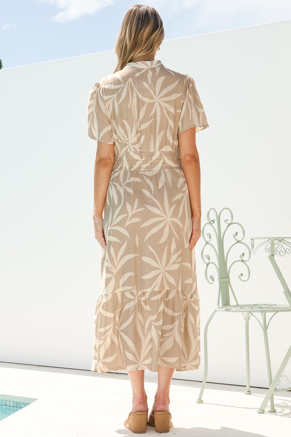 Back view of a woman wearing the Paloma Midi Dress in Gretchen Print, showing the collar, cap sleeves and tiered A-line skirt of the beige and cream leaf-print midi, with tan sandals on a sunny white terrace