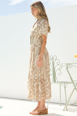 Side profile of a woman wearing the Paloma Midi Dress in Gretchen Print, showing the cap sleeve, cinched waist and flowing tiered skirt of the beige leaf-print midi, with tan sandals on a sunny white terrace