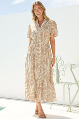 A smiling woman mid-step in the Paloma Midi Dress in Gretchen Print, a flowy beige leaf-print button-down midi with a split mandarin collar and cap sleeves, on a bright white terrace with a garden chair