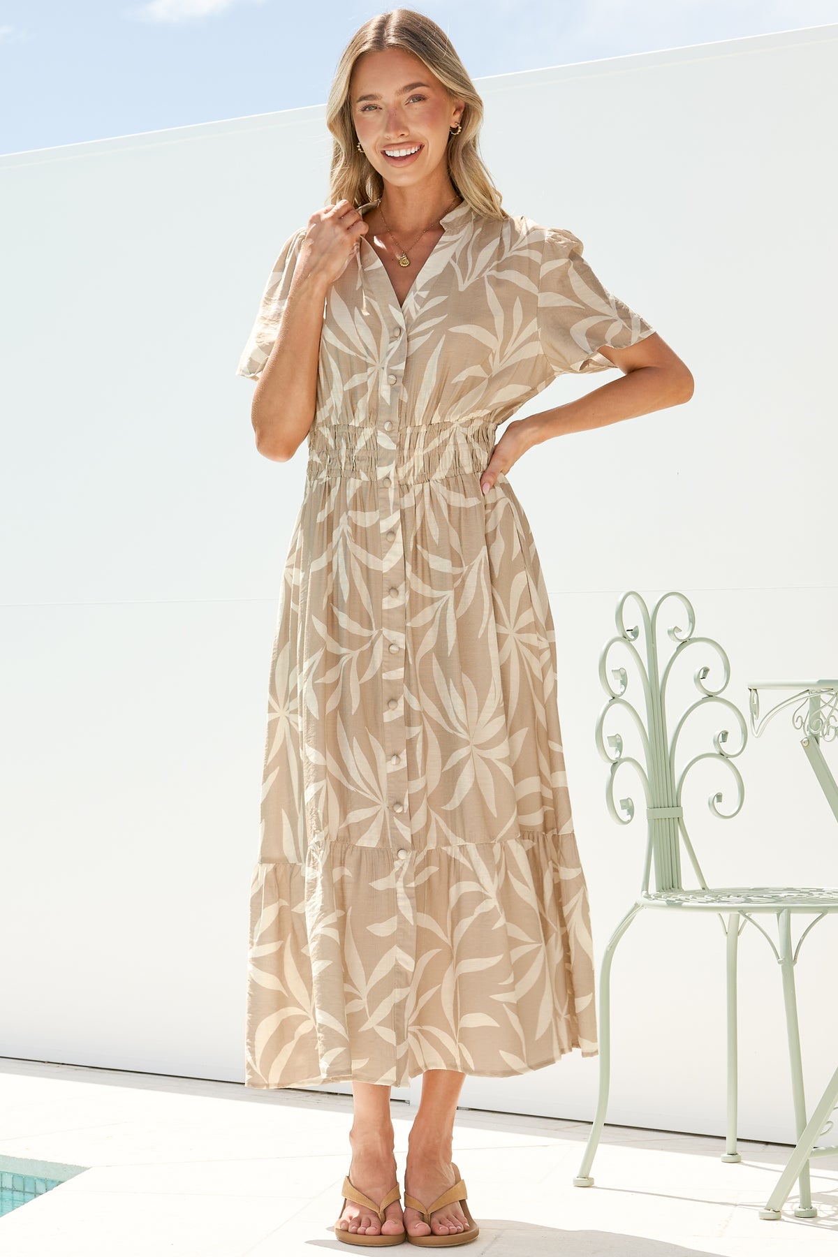 A smiling woman with one hand on her hip wearing the Paloma Midi Dress in Gretchen Print, showing the split mandarin collar, button front and tiered hem of the beige leaf-print midi, on a bright white terrace with a garden chair