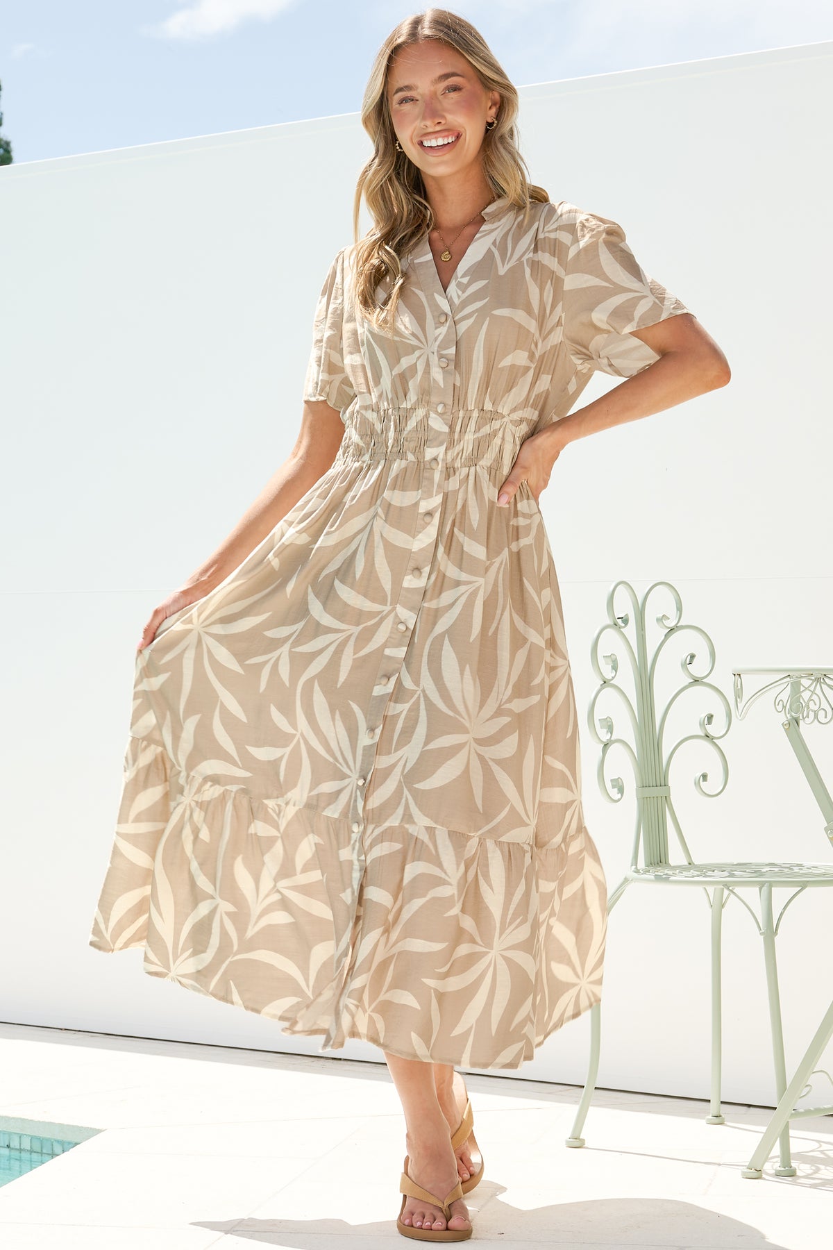 A smiling woman holding out the skirt of the Paloma Midi Dress in Gretchen Print, showing the full A-line shape and beige and cream leaf print, with a hand on her hip on a sunny white terrace