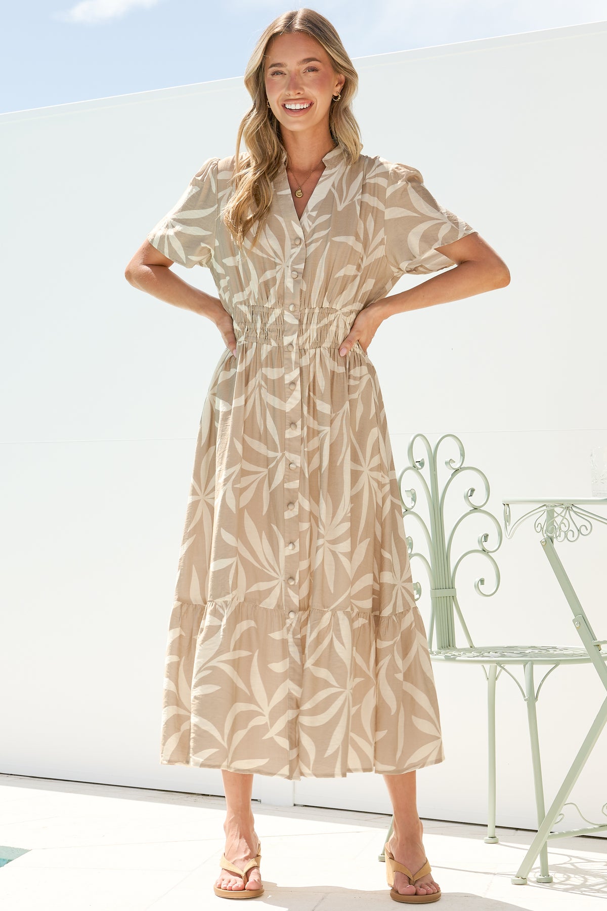 A smiling woman with both hands on her waist wearing the Paloma Midi Dress in Gretchen Print, showing the button front, cap sleeves and tiered A-line skirt of the beige leaf-print midi, on a bright white terrace