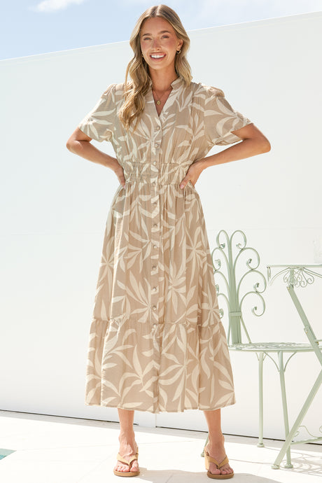 A smiling woman with both hands on her waist wearing the Paloma Midi Dress in Gretchen Print, showing the button front, cap sleeves and tiered A-line skirt of the beige leaf-print midi, on a bright white terrace