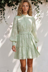 Woman wearing a light green dress standing against a white wall with shadows.