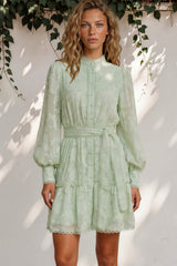 Woman wearing a light green dress standing against a white wall with foliage.