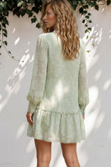 Woman wearing a light green dress standing against a white wall with greenery.