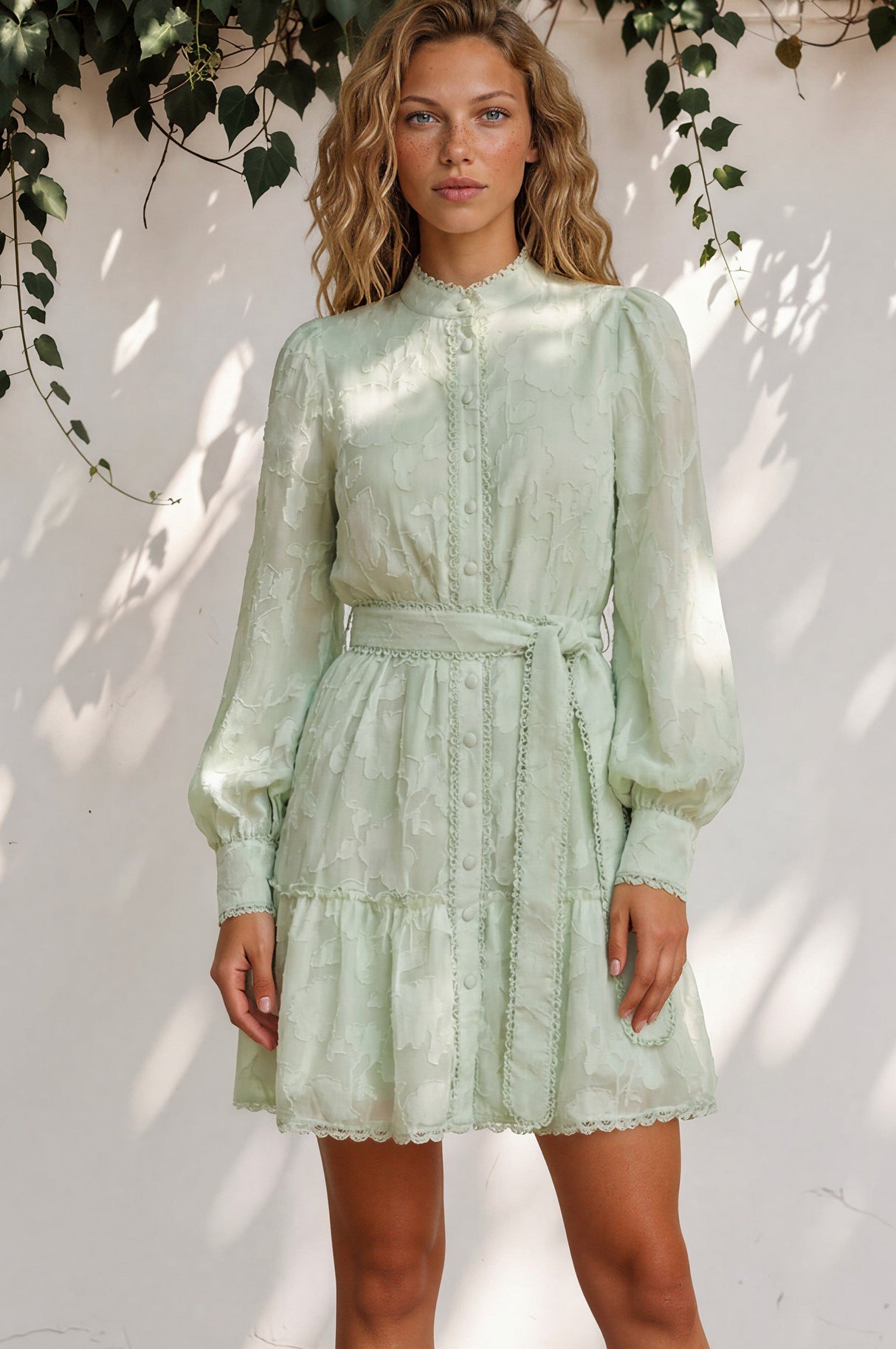 Woman wearing a light green dress standing against a white wall with leafy branches above.