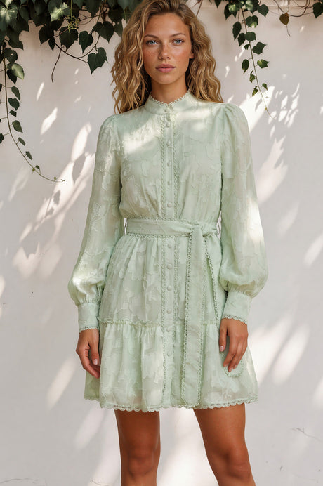 Woman wearing a light green dress standing against a white wall with leafy branches above.