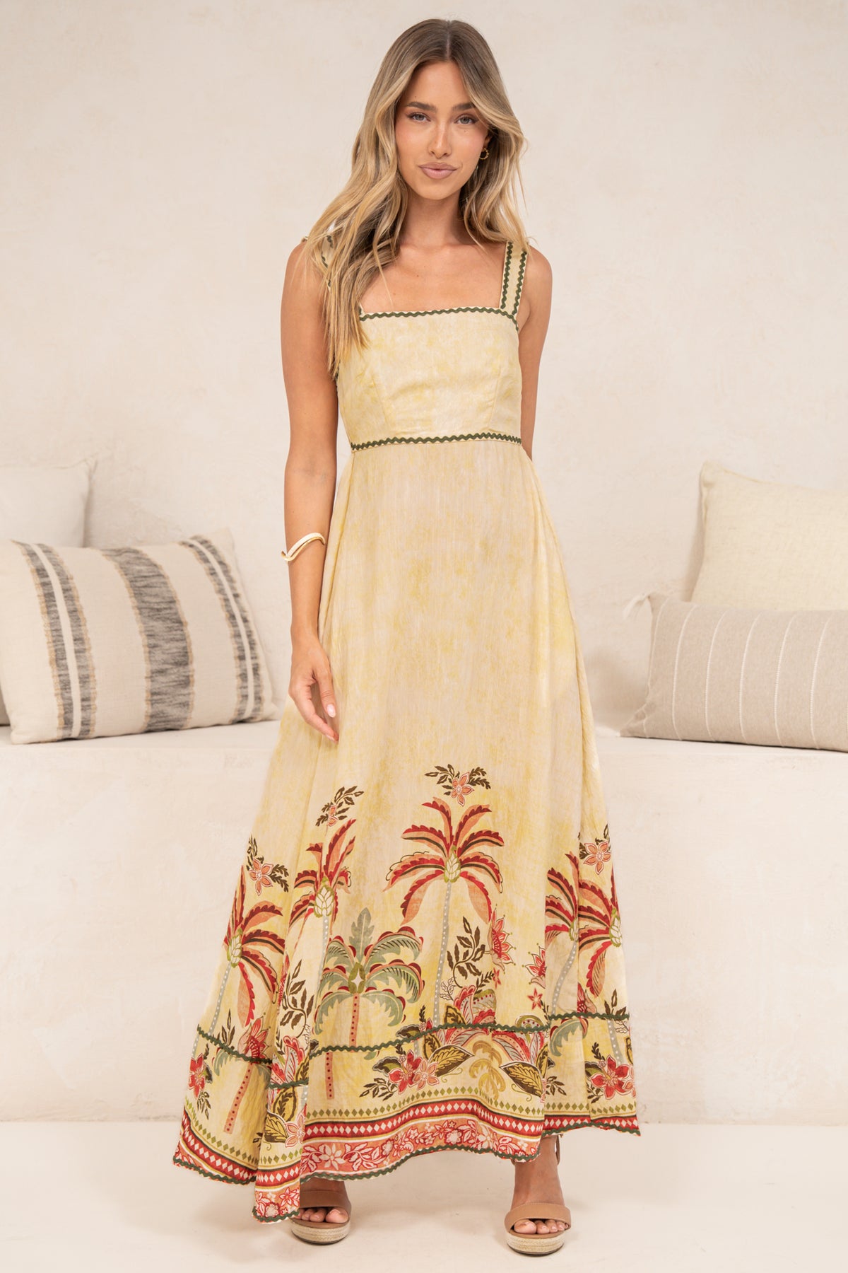 A woman standing front-on in the Poppy Maxi Dress in Palmina Print, showing the full length of the butter yellow A-line sun dress with thick straps, ric rac trims and palm tree border print, styled with tan sandals in front of a cream sofa
