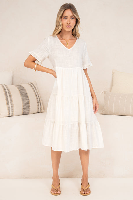A woman wearing the Porcelaine Midi Dress in white, a tiered V-neck midi dress with short frill-trimmed sleeves, styled with tan sandals, standing in front of a cream sofa with striped and neutral cushions