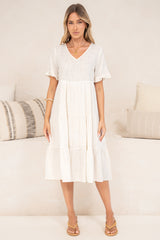 A woman standing front-on in the Porcelaine Midi Dress in white with arms relaxed at her sides, showing the full tiered midi length, V-neckline and frill-trimmed short sleeves, with tan sandals in front of a cream sofa with neutral cushions