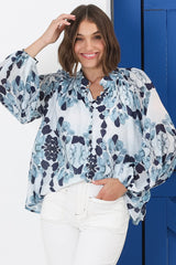 A woman wearing a blue floral button-down blouse with a high mandarin collar and balloon sleeves, paired with white trousers.