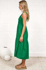 A woman in a flowing green midi dress featuring a shirred bodice and tiered skirt, posed against a white painted brick wall.