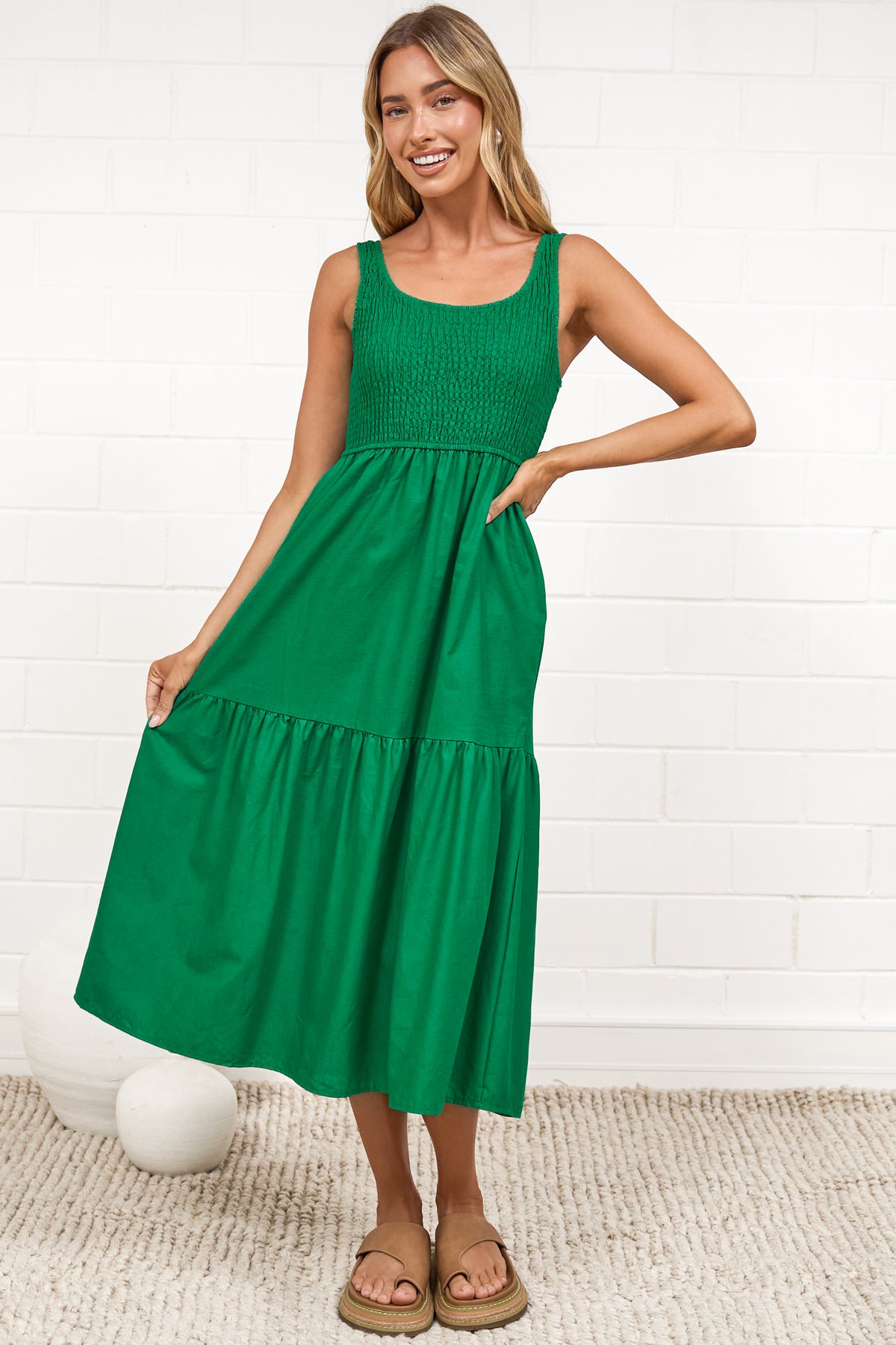 A woman in a flowing green midi dress featuring a shirred bodice and tiered skirt, posed against a white painted brick wall.