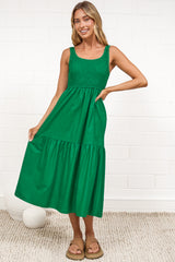 A woman in a flowing green midi dress featuring a shirred bodice and tiered skirt, posed against a white painted brick wall.