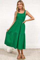 A woman in a flowing green midi dress featuring a shirred bodice and tiered skirt, posed against a white painted brick wall.