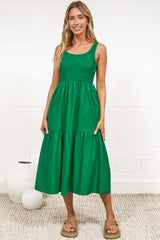 A woman in a flowing green midi dress featuring a shirred bodice and tiered skirt, posed against a white painted brick wall.