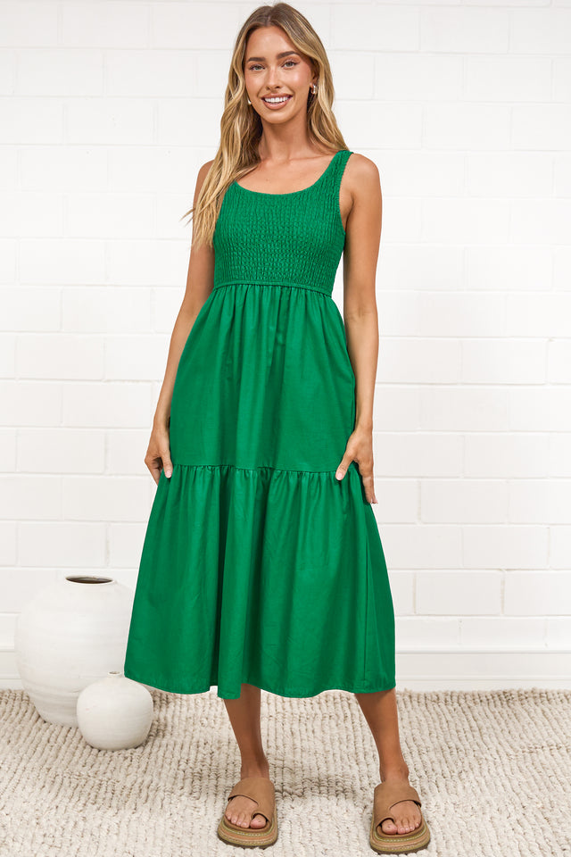 A woman in a flowing green midi dress featuring a shirred bodice and tiered skirt, posed against a white painted brick wall.