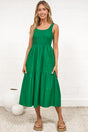 A woman in a flowing green midi dress featuring a shirred bodice and tiered skirt, posed against a white painted brick wall.