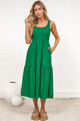 A woman in a flowing green midi dress featuring a shirred bodice and tiered skirt, posed against a white painted brick wall.