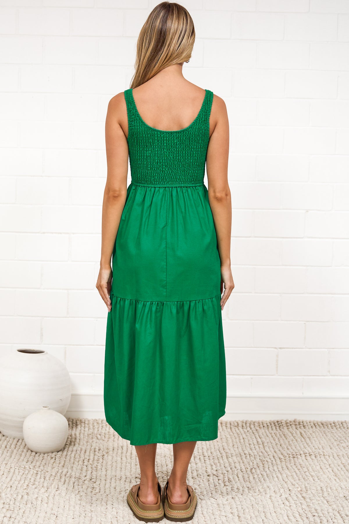 A woman in a flowing green midi dress featuring a shirred bodice and tiered skirt, posed against a white painted brick wall.