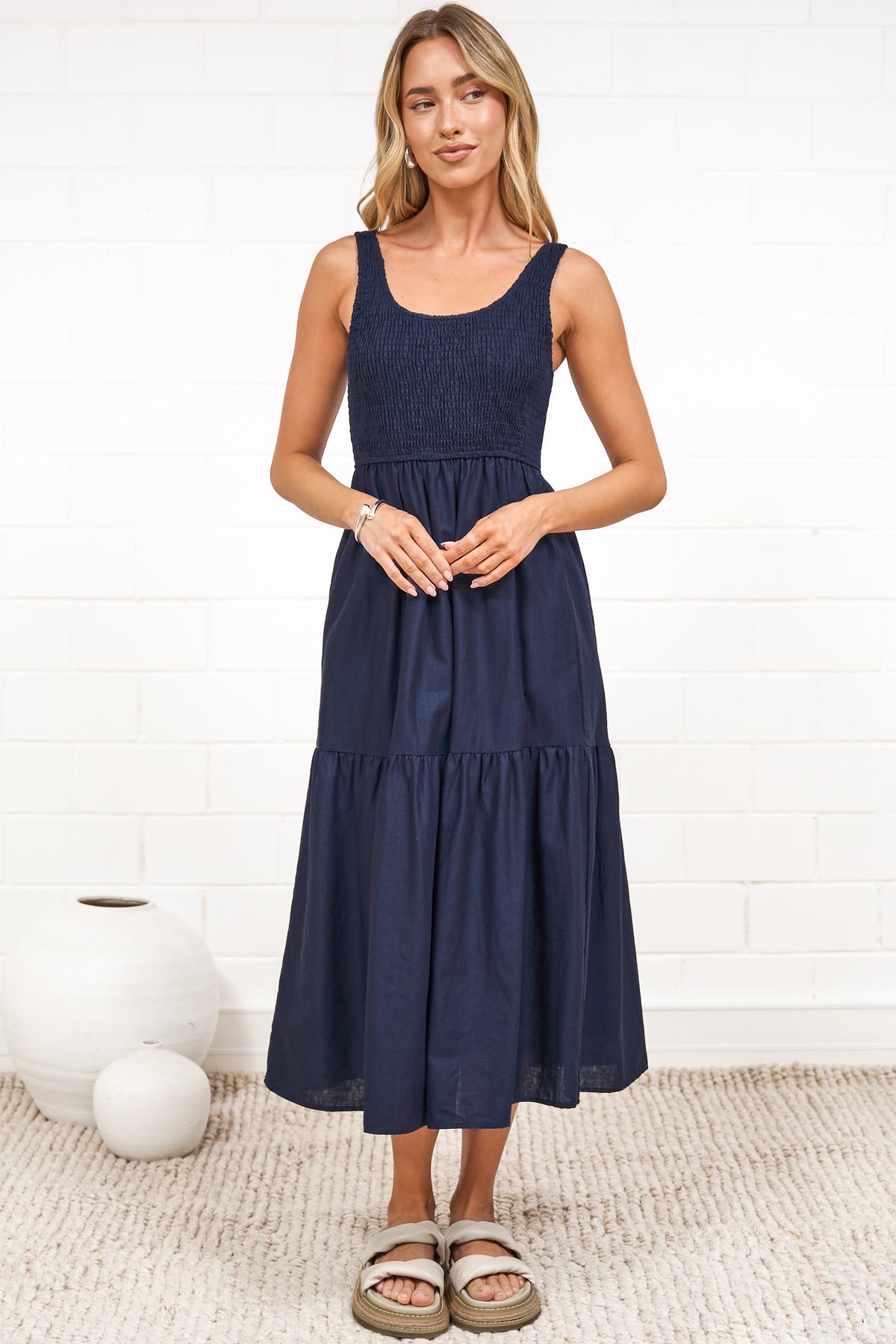A woman in a rust midi dress with thick straps, a shirred bodice, and tiered skirt, posed against a white painted brick wall.