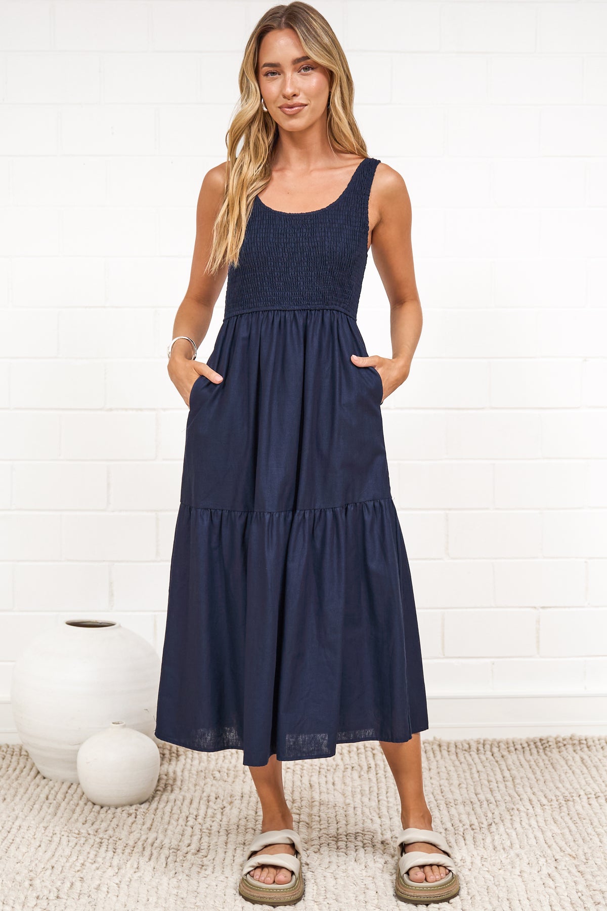 A woman in a rust midi dress with thick straps, a shirred bodice, and tiered skirt, posed against a white painted brick wall.