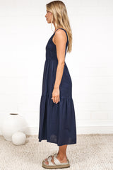 A woman in a rust midi dress with thick straps, a shirred bodice, and tiered skirt, posed against a white painted brick wall.