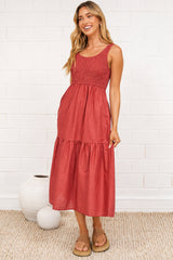 A woman in a rust midi dress with thick straps, a shirred bodice, and tiered skirt, posed against a white painted brick wall.